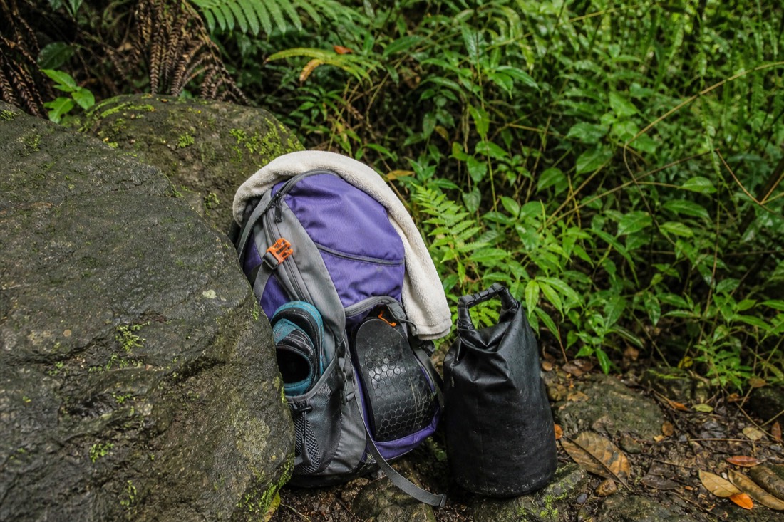 Daypack open on a wooden lanai bench with a rolled quick-dry towel, worn water shoes, a rolled dry bag and a steel water bottle, soft late-morning light and wet greenery behind